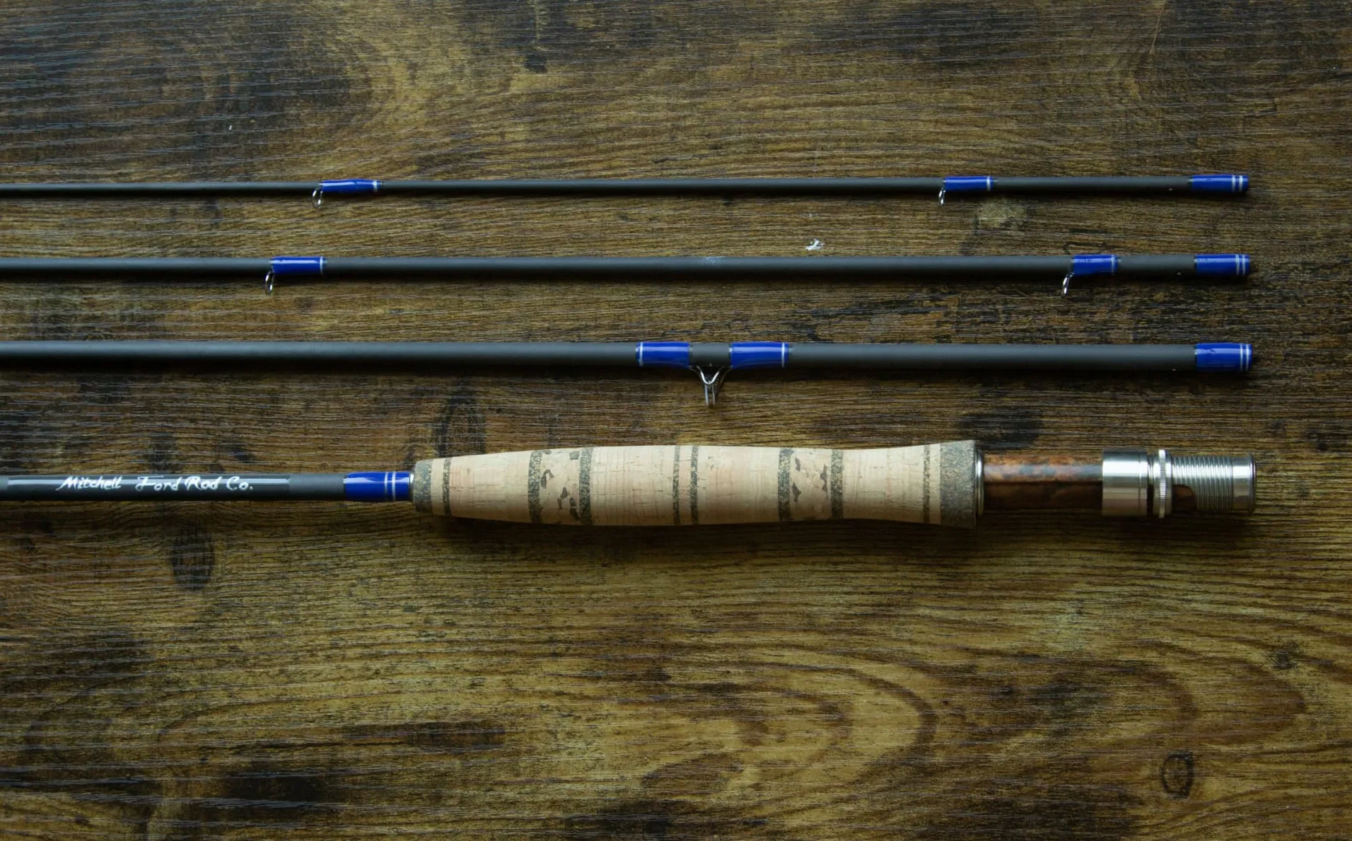 Four-piece Mitchell Ford fly rod with blue wraps, cork grip and burl reel seat on a wooden table